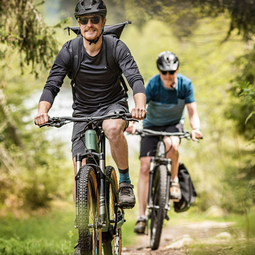 Zwei Personen fahren gemeinsam mit E-Mountainbikes durch einen sonnigen Waldpfad.
