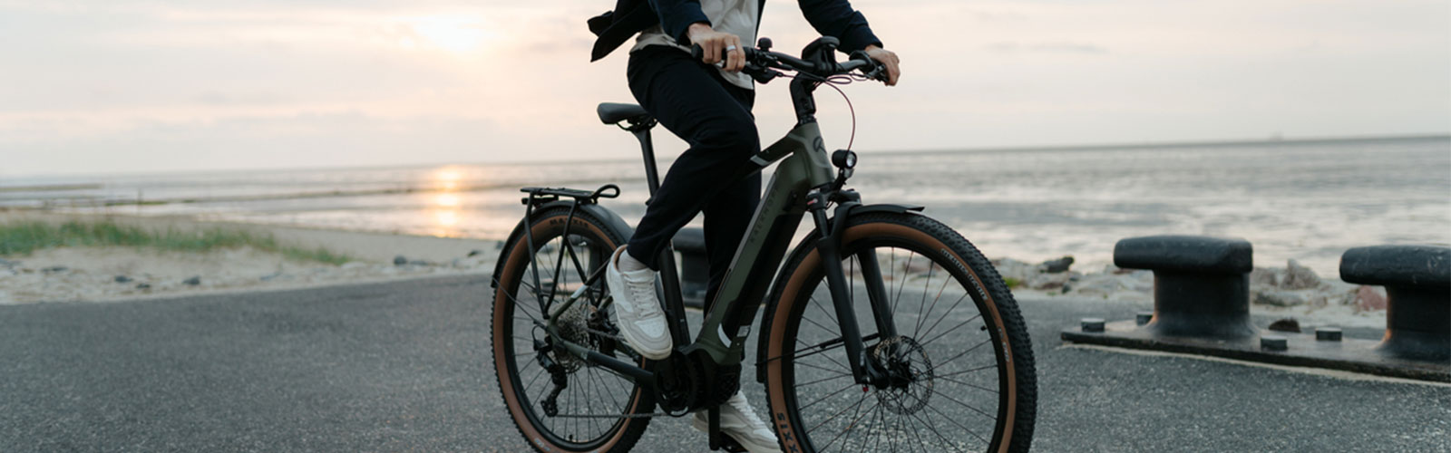 Person mit E-Bike am Meer bei Sonnenuntergang – modernes Citybike mit braunen Reifen auf einer Promenade.