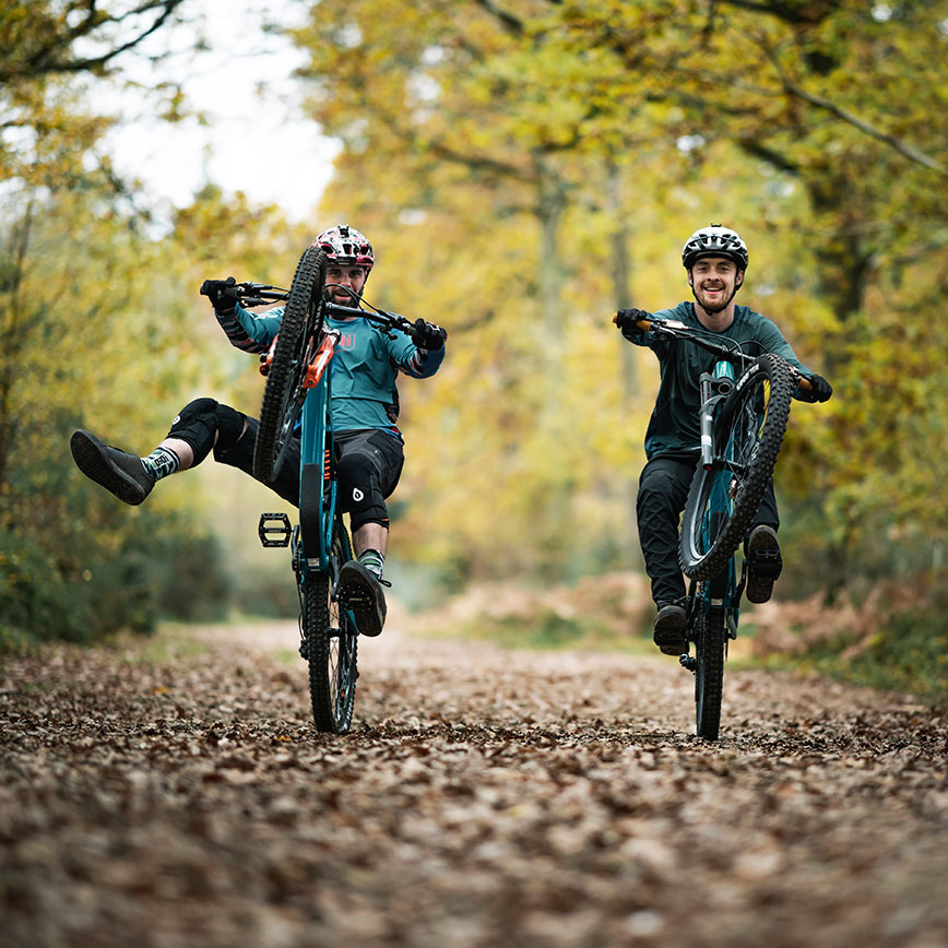 Zwei Mountainbiker fahren Wheelies auf einem herbstlich bedeckten Waldweg – Spaß und Action beim Trailfahren.